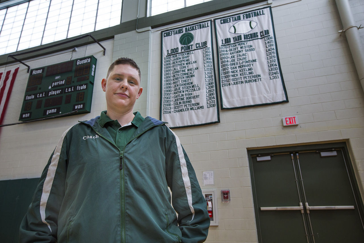 Stephen Anderson stands in front of the Ponaganset High School Chieftains 1,000 point club banner for basketball players where he is recognized for his achievement as Jen Dandrow..He has asked the school to change his name on the banner. (Jesse Costa/WBUR)